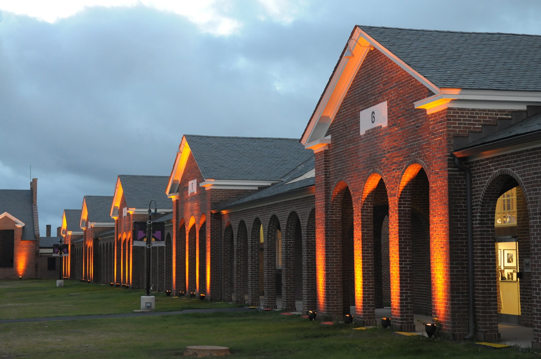 A row of brick buildings with arched openings, illuminated by orange lights along the exterior at dusk or evening, with a cloudy sky overhead.
