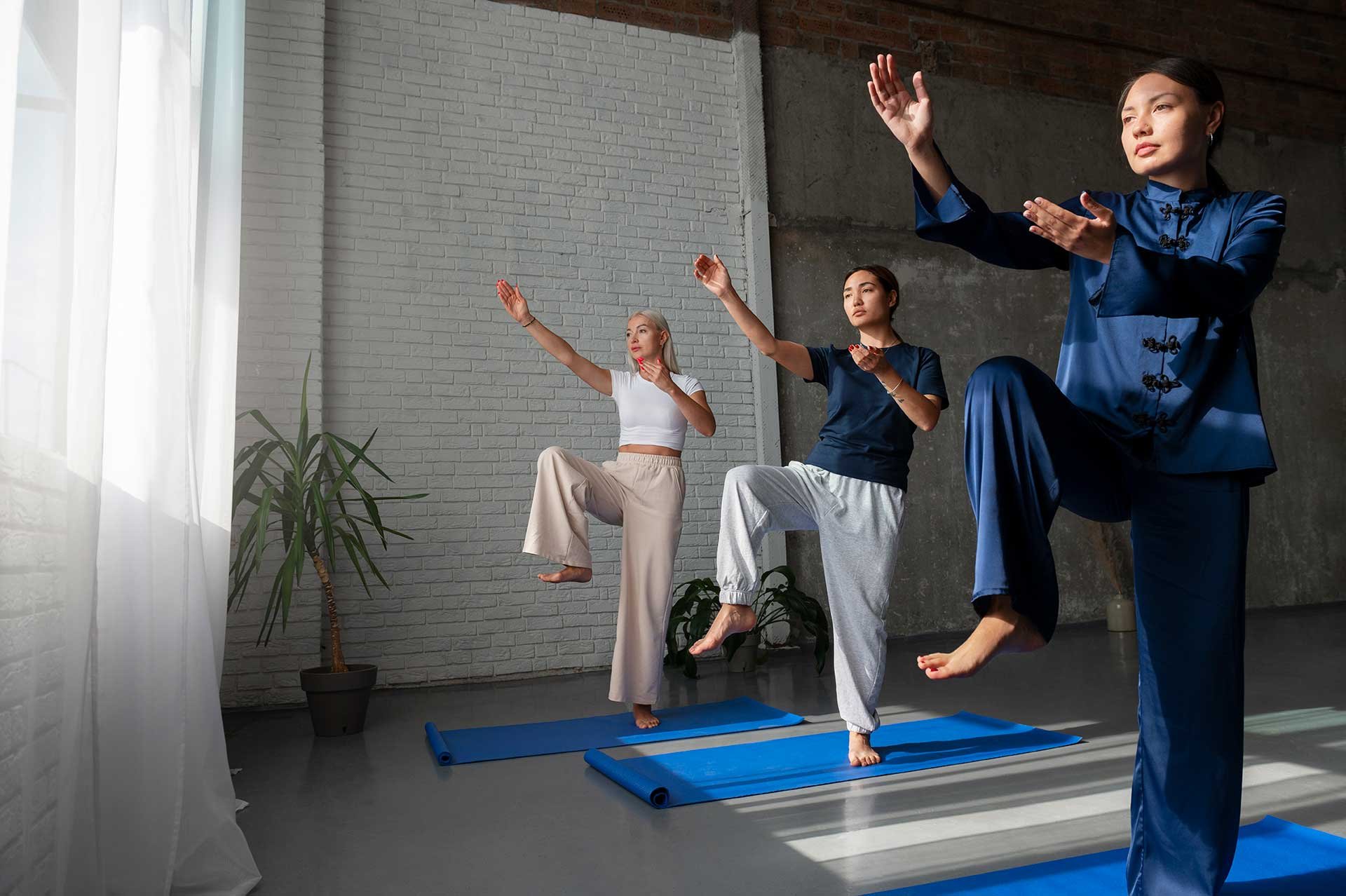Three women practicing yoga indoors on blue mats, balancing on one leg with arms raised, in a room with white brick wall, large window with curtains, and potted plants.
