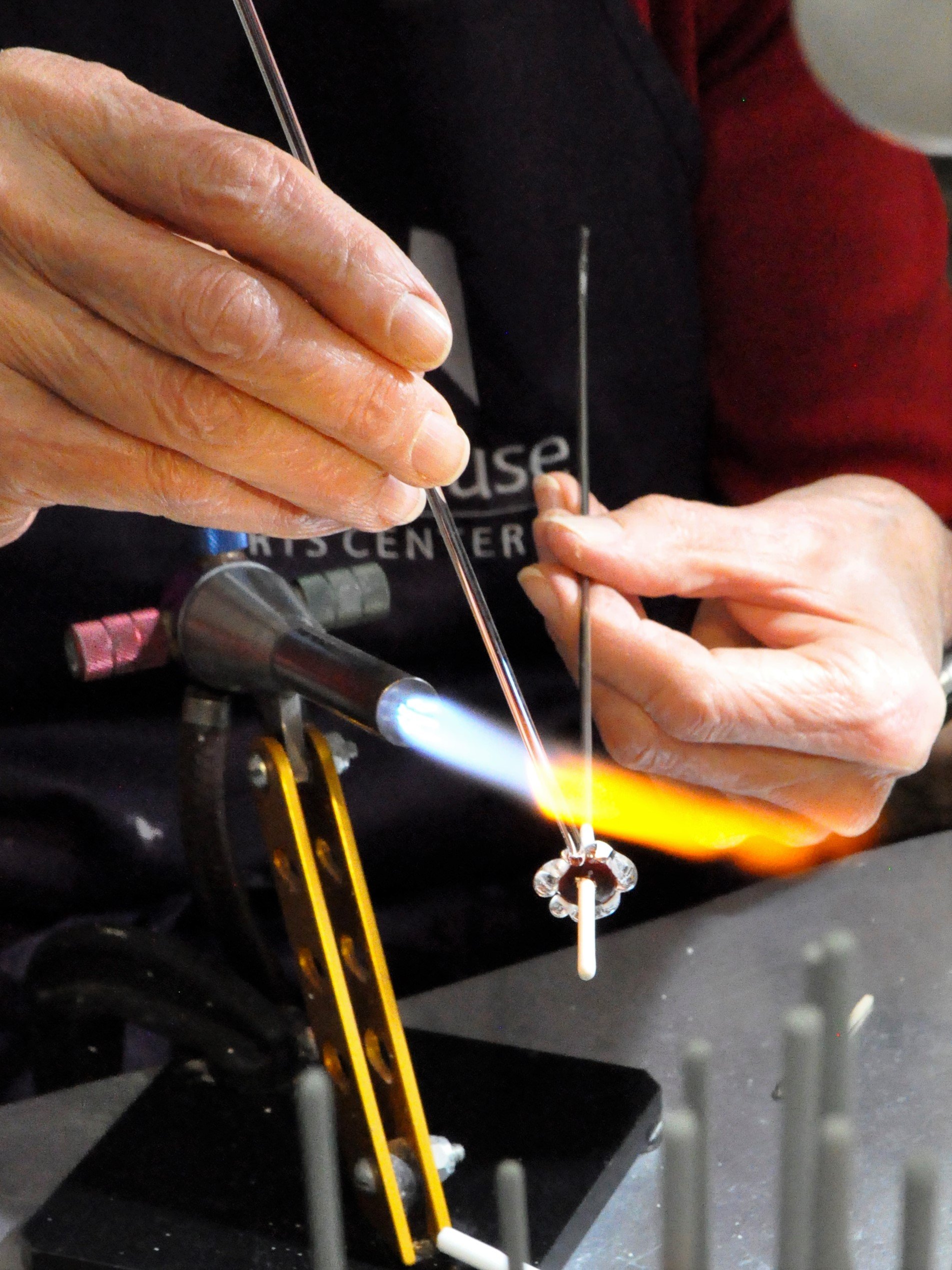 Person using a blowtorch to work on a glass piece, with hands holding tools, and a small flame and glowing orange heating element visible.