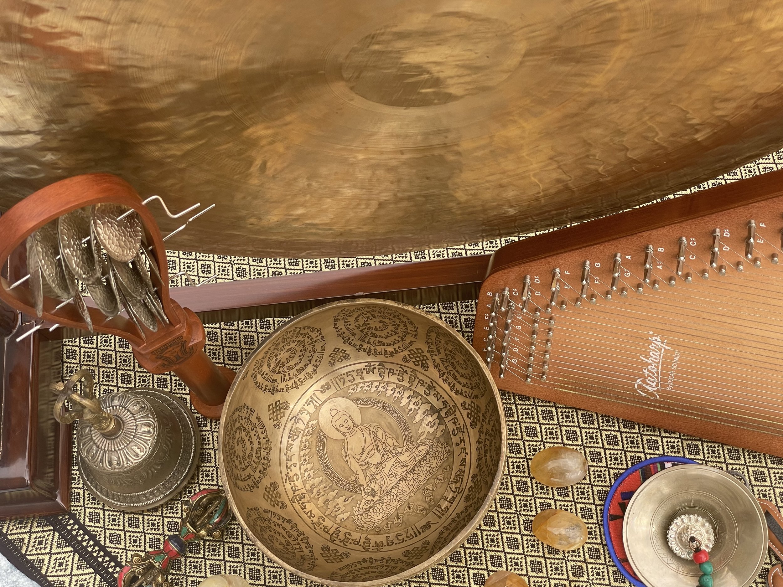Collection of cultural and musical objects including a hanging set of bells, a large decorated bowl with Buddha and script, a wooden musical instrument with metal keys, a brass bell, and small stones on patterned cloth