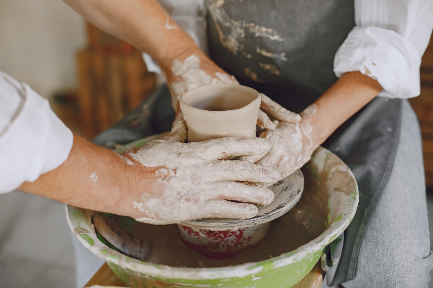 Two people shaping a clay pot on a pottery wheel, with one person guiding the hands of the other. Both have chalky white clay on their hands and arms.