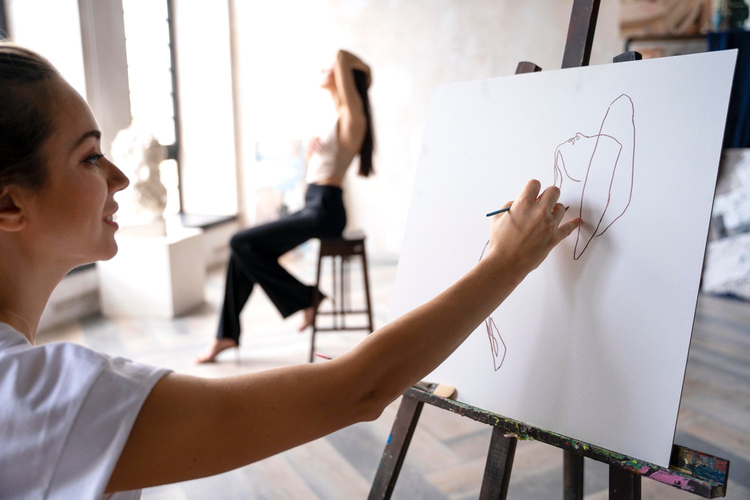 A woman drawing a line art portrait on a white canvas in an art studio, with another woman sitting on a stool in the background.
