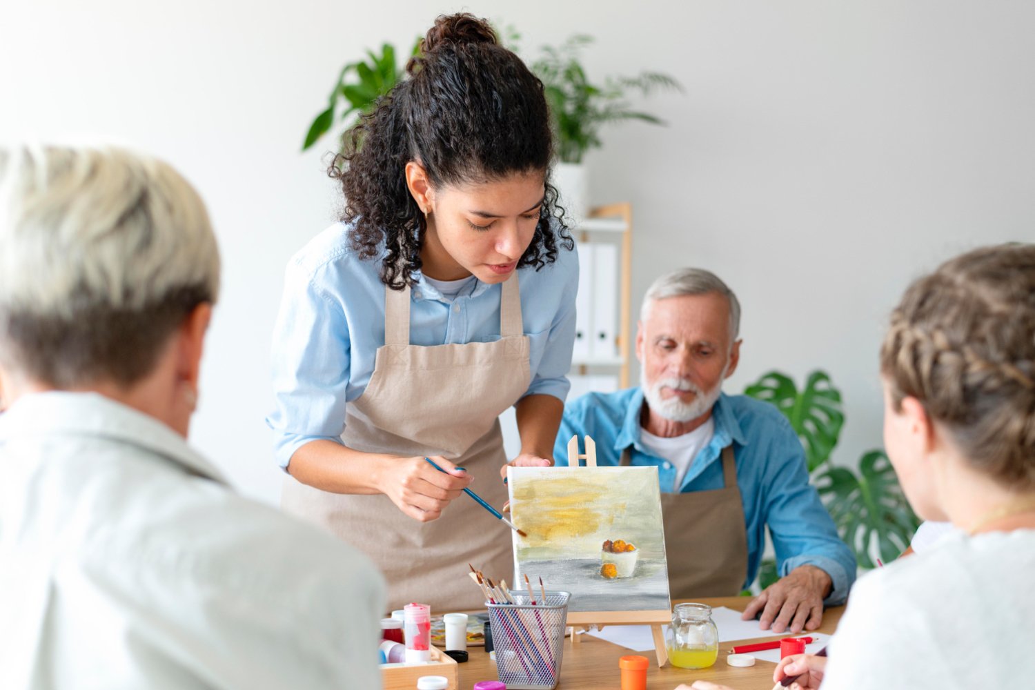 A young woman is painting a still life on an easel while three older adults watch. The room has plants and shelves with binders.