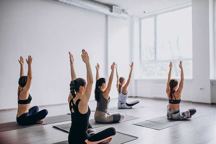 Group of women participating in a yoga or meditation class, sitting on yoga mats in a bright, airy studio with large windows, raising their arms above their heads.